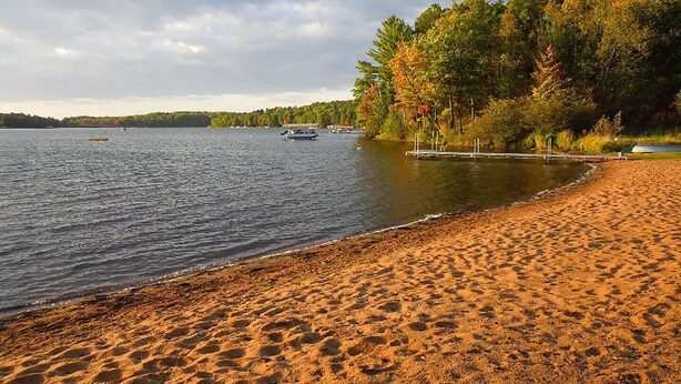 A sandy beach with a calm lake, boats, and trees with autumn foliage. - Sandys Beach House (Hayward)