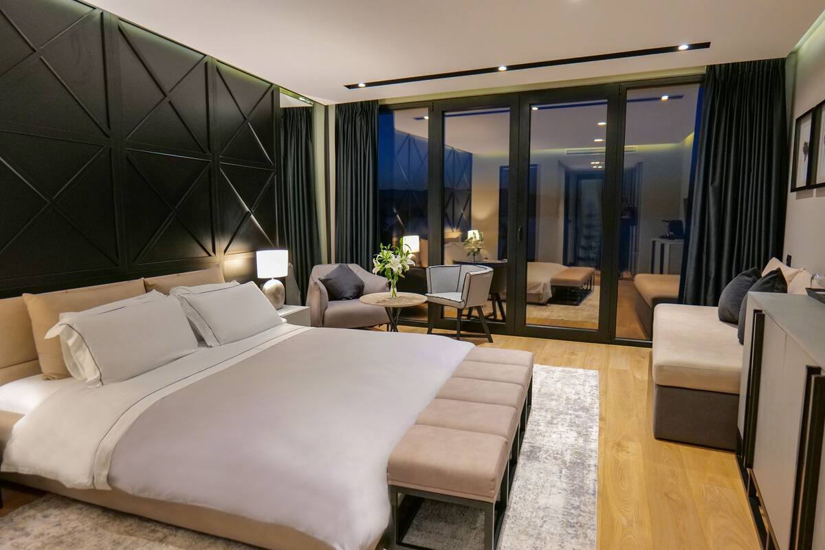 Quarto de hotel moderno com uma cama grande, uma escrivaninha, uma cadeira e vista através de uma porta de vidro.
