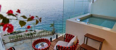 A balcony with a view of the sea, a hot tub, and red and black wicker chairs.