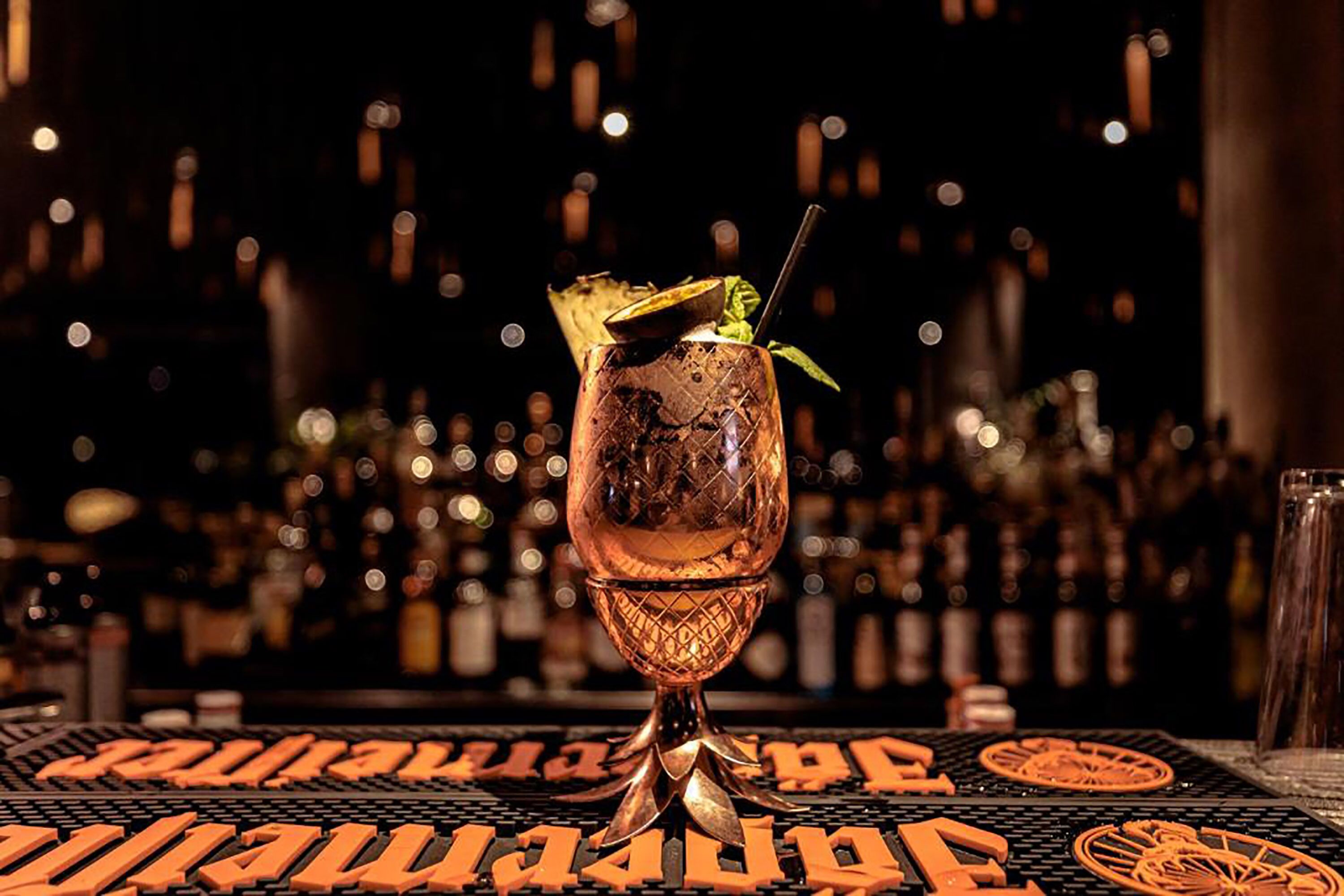 Un cocktail servi dans un verre en cuivre, posé sur le comptoir d’un bar, avec un arrière-plan flou composé de bouteilles.