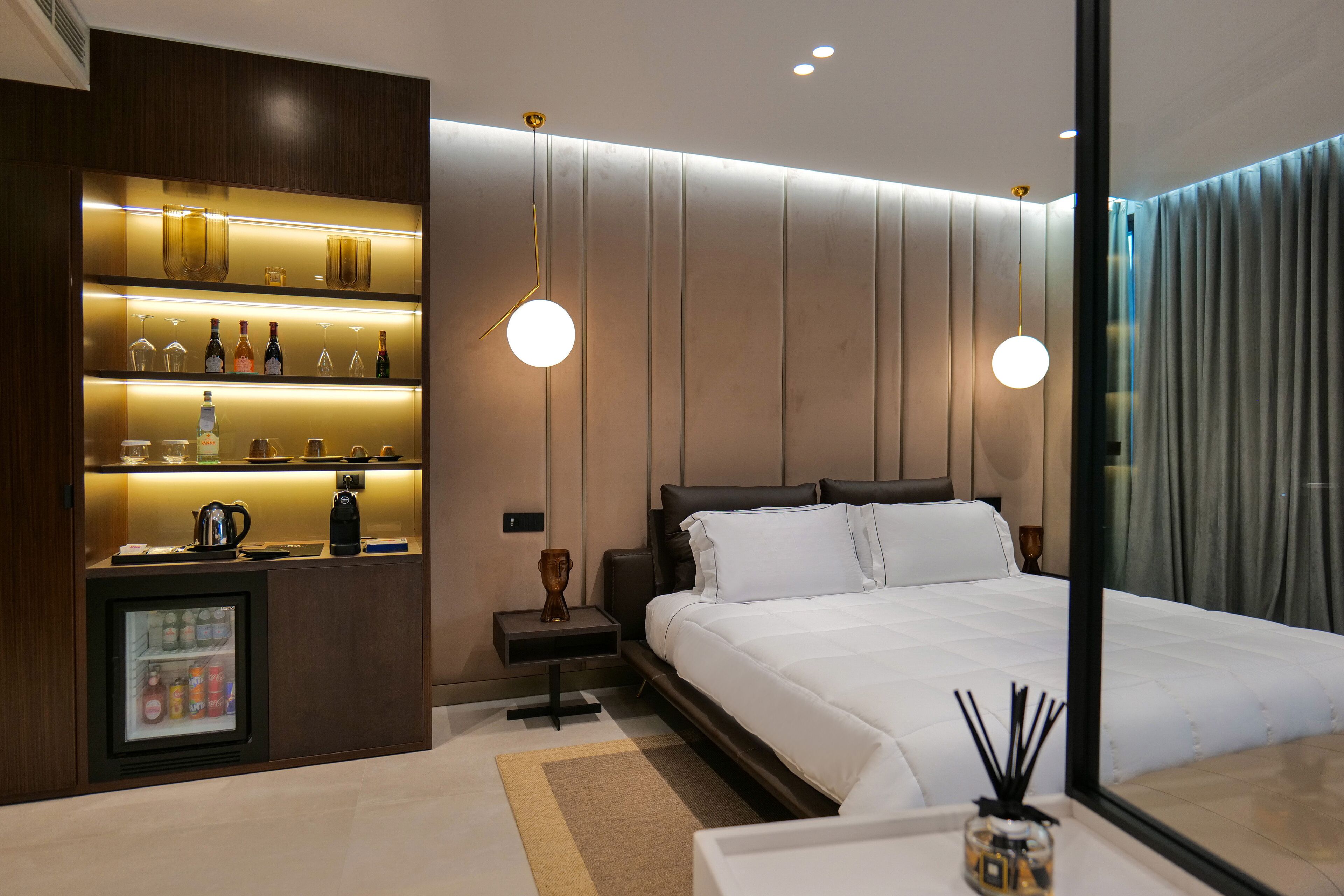 A modern hotel room with a large bed, a minibar, and a glass-enclosed refrigerator.