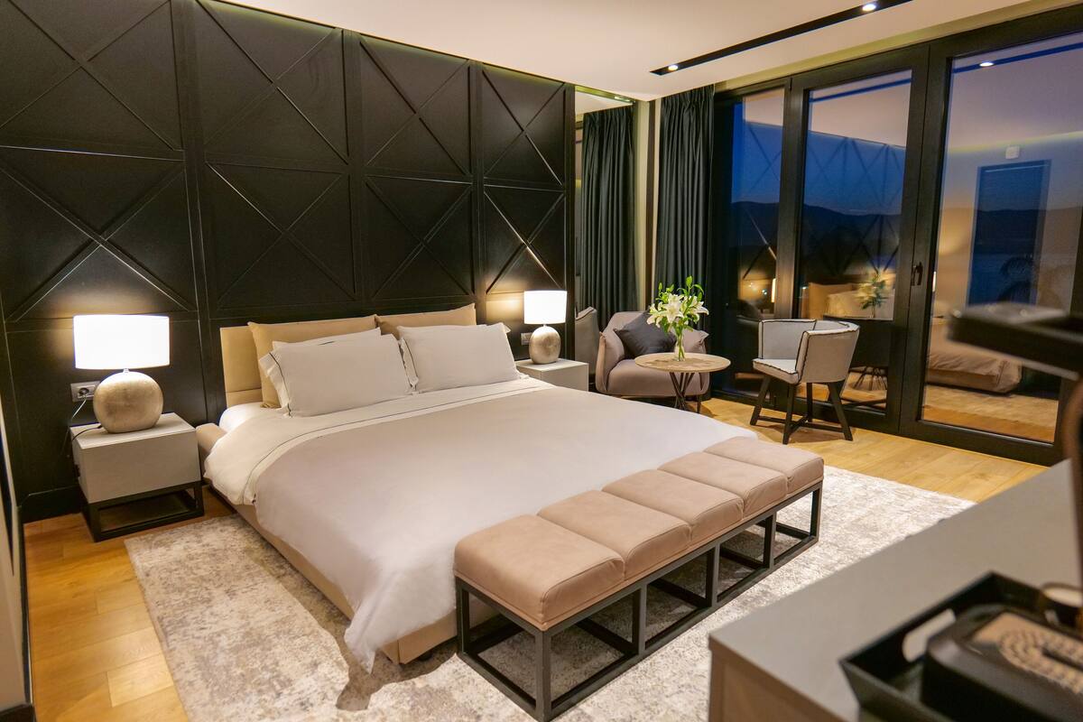 Quarto de hotel moderno com uma cama grande, mesas de cabeceira, um banco, uma pequena mesa com cadeiras e vista para a paisagem urbana através de amplas janelas.