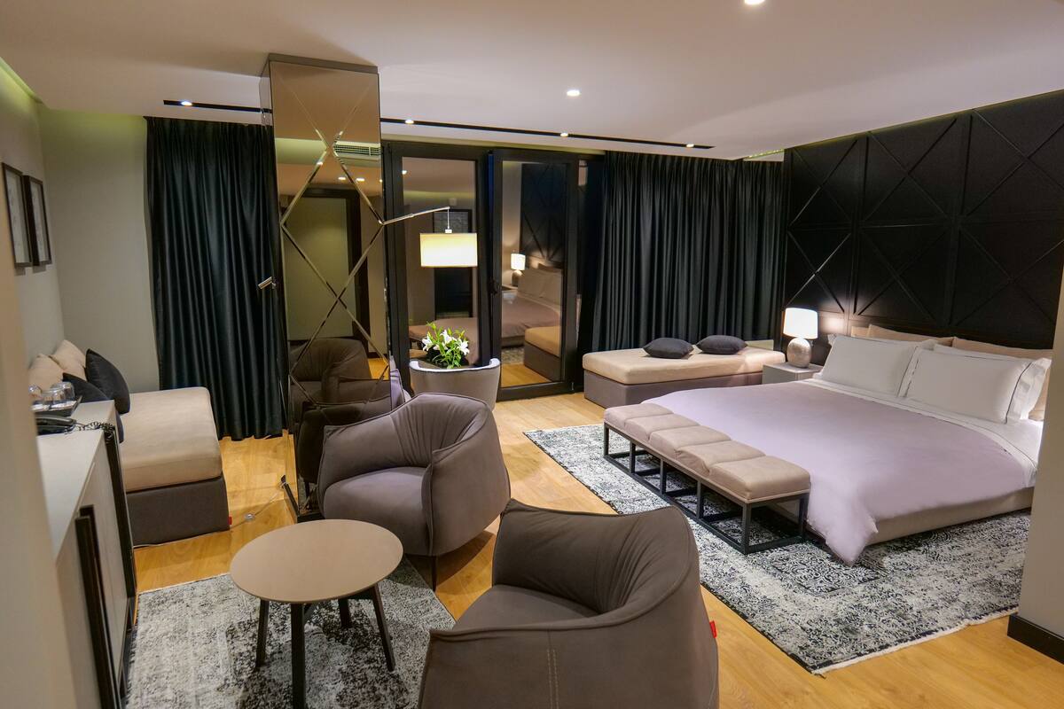 Quarto de hotel moderno com uma cama grande, um sofá, uma mesinha e uma cadeira. Há uma escrivaninha com uma luminária e uma planta. O quarto possui uma janela ampla com cortinas.