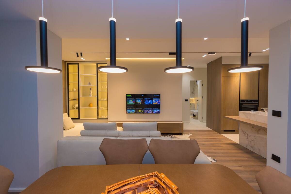 Uma sala de estar moderna com área de jantar, TV fixada na parede e cozinha visível por uma porta aberta.