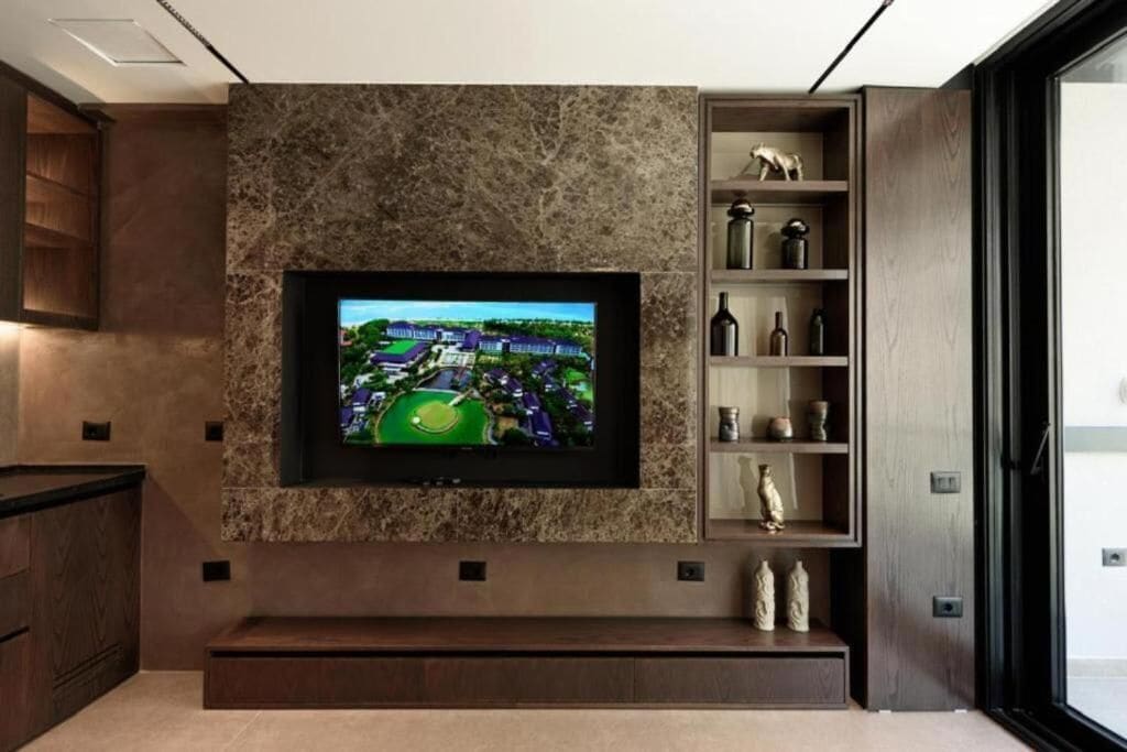 A modern living room with a flat-screen TV mounted on a textured wall, a built-in bench, and a wooden shelving unit with decorative items.