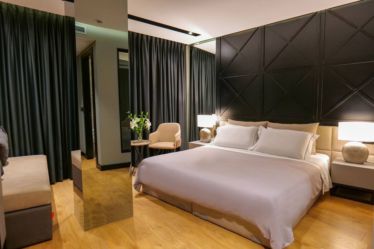 Quarto de hotel moderno com uma cama grande, área de estar com uma cadeira e uma mesa pequena, e um painel decorativo na parede.