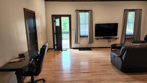 A living room with a wooden floor, a black sofa, a TV on a stand, a desk with a chair, and a door leading outside.