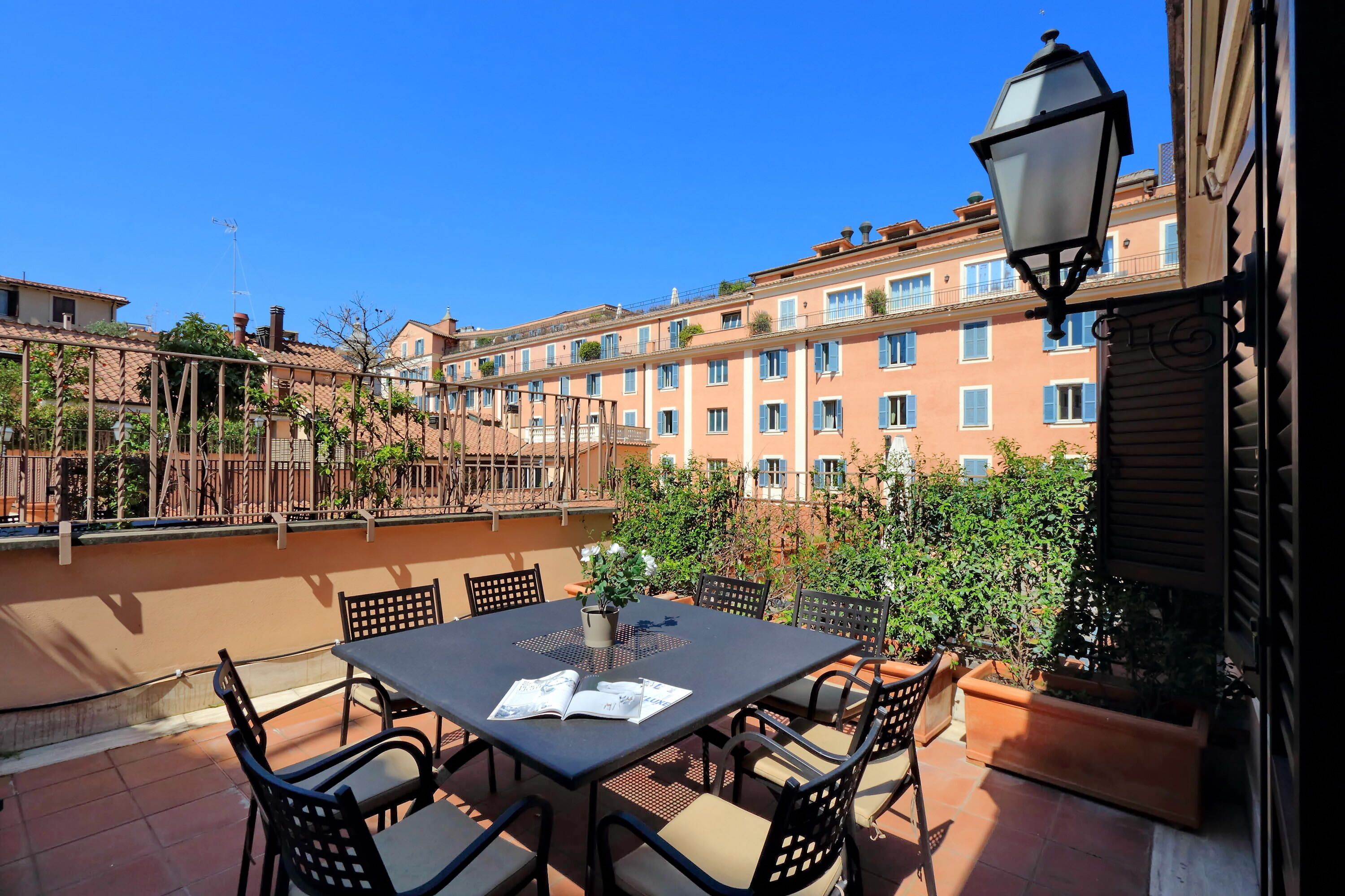 A terrace with a table and chairs, a lamp, and a view of a building with balconies and windows.