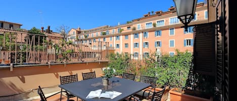 A terrace with a table and chairs, a lamp, and a view of a building with balconies and windows.