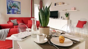 En moderne stue med en rød sofa, et sofabord med en muffin, en avis, en kande vand og en potteplante.