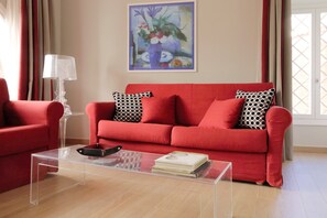 Un canapé rouge agrémenté de coussins à motifs noirs et blancs, une table basse en verre transparent sur laquelle reposent un livre et un plateau noir, et un tableau représentant des fleurs accroché au mur.