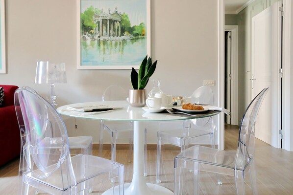 A dining area with a round glass table, clear chairs, a potted plant, a plate of food, and a painting on the wall.