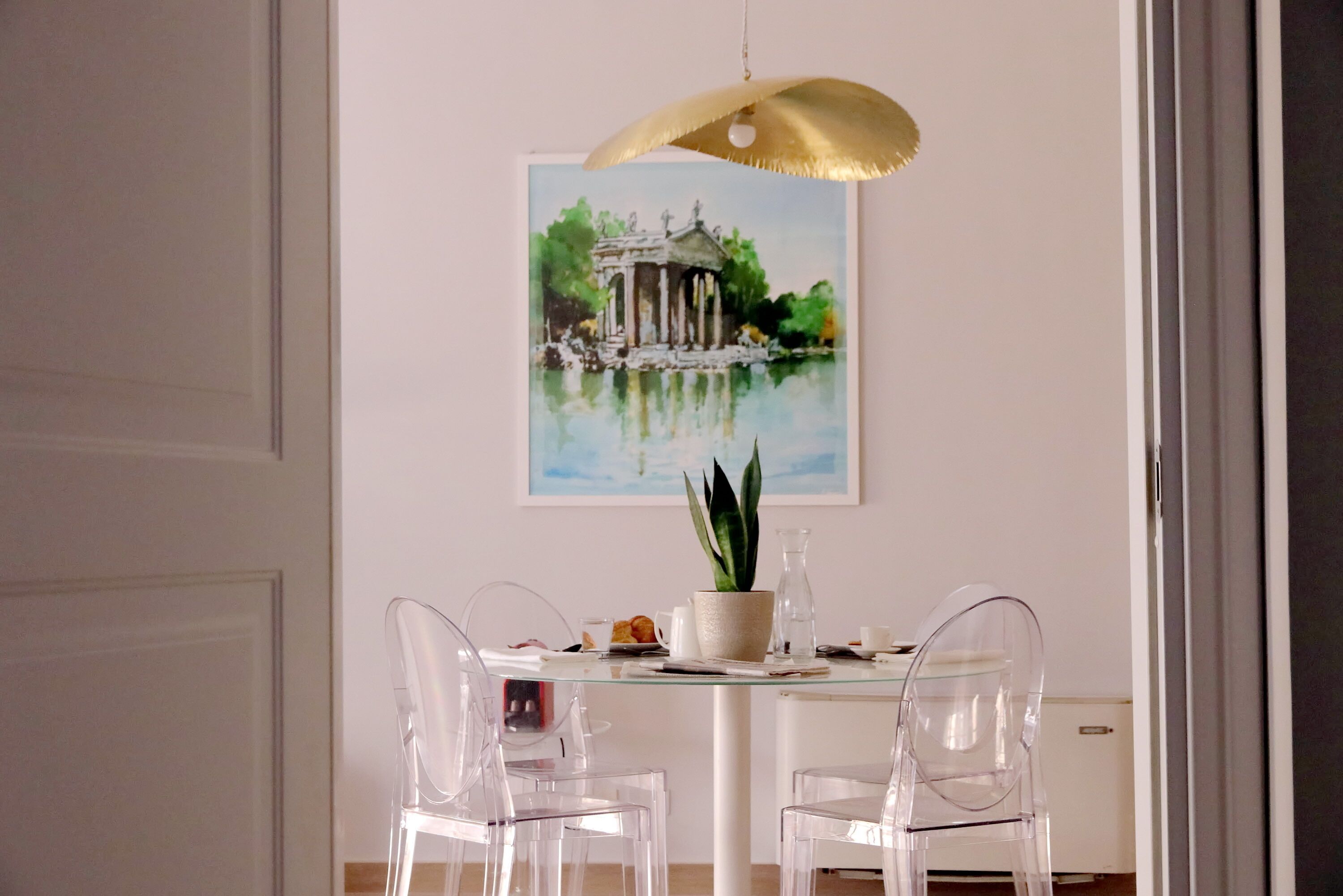 A dining area with a round table, clear chairs, a potted plant, and a painting of a classical building.