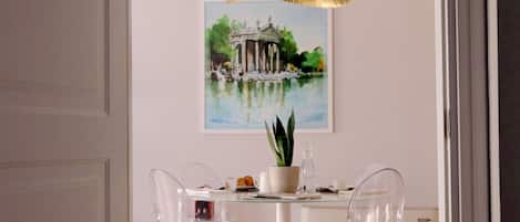 A dining area with a round table, clear chairs, a potted plant, and a painting of a classical building.