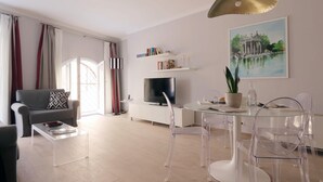 Una sala de estar moderna con un sofá gris, una mesa de centro de cristal, un televisor y una zona de comedor con sillas transparentes.
