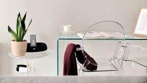 Een moderne kamer met een glazen tafel, een potplant, een witte kop, een zwart oplaadstation en een transparante stoel.