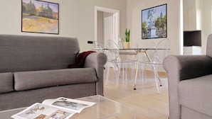 Een moderne woonkamer met een grijze bank, een glazen salontafel met tijdschriften, en een eethoek met transparante stoelen en een witte tafel.