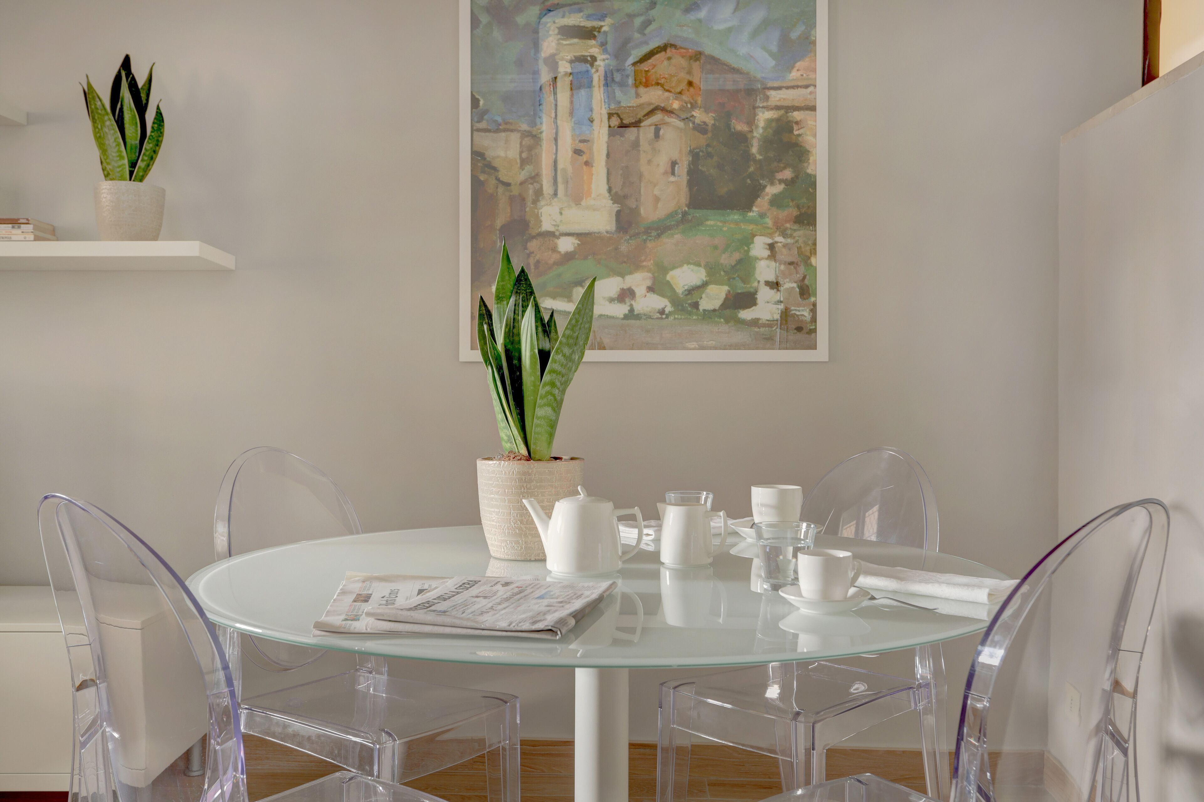 A modern dining area with a glass table, clear chairs, a white pitcher, cups, and a newspaper. There are two potted plants on a shelf above the table.