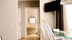 A hotel room with a wooden floor, a glass table, a mounted TV, and a bathroom with a sink and mirror. - Piazzetta Margutta - T1 (Rome)