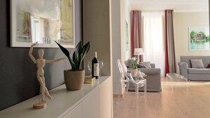 A modern living room with a wooden mannequin, a potted plant, and a wine bottle on a white console table. There are clear chairs, a grey sofa, and a glass coffee table. - Piazzetta Margutta - T1 (Rome)