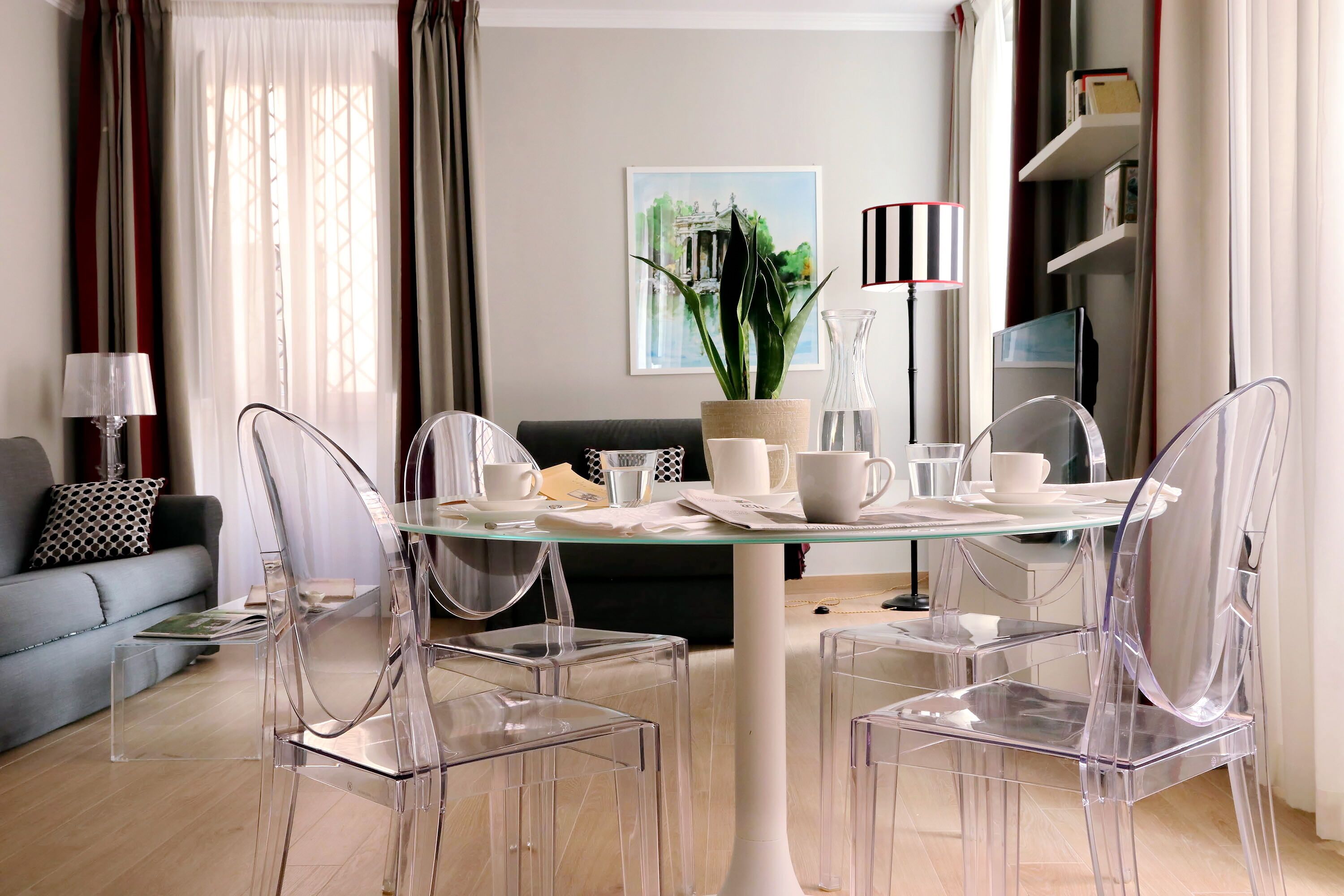A modern living room with a glass dining table, clear chairs, a grey sofa, and a bookshelf.