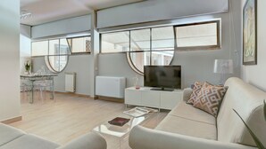 A modern living room with a sofa, coffee table, and a TV on a stand. There is a dining area with a table and chairs, and a window with a view of the outside. - Piazzetta Margutta - A1 (Rome)