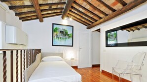 A bedroom with a bed, wooden ceiling beams, a framed picture on the wall, and a clear plastic chair. - Piazzetta Margutta - B6 (Rome)
