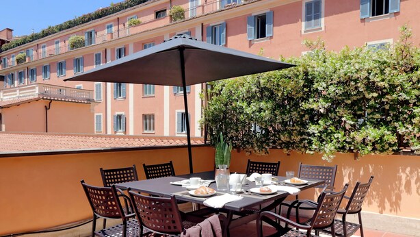 Una terraza con mesa lista para dos personas, rodeada de sillas, una sombrilla y una planta en maceta.