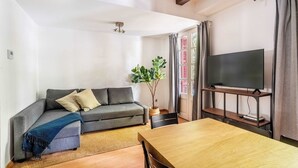 A living room with a grey sofa, a wooden dining table, a flat-screen TV on a stand, and a potted plant. - Pl Universitat Comfortable 3BD with private patio (Barcelona)