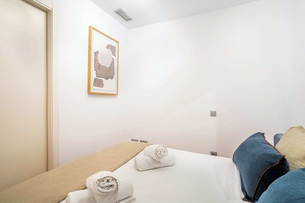 A hotel room with a bed, a beige pillow, a white pillow, and a blue pillow. There is a framed picture on the wall.