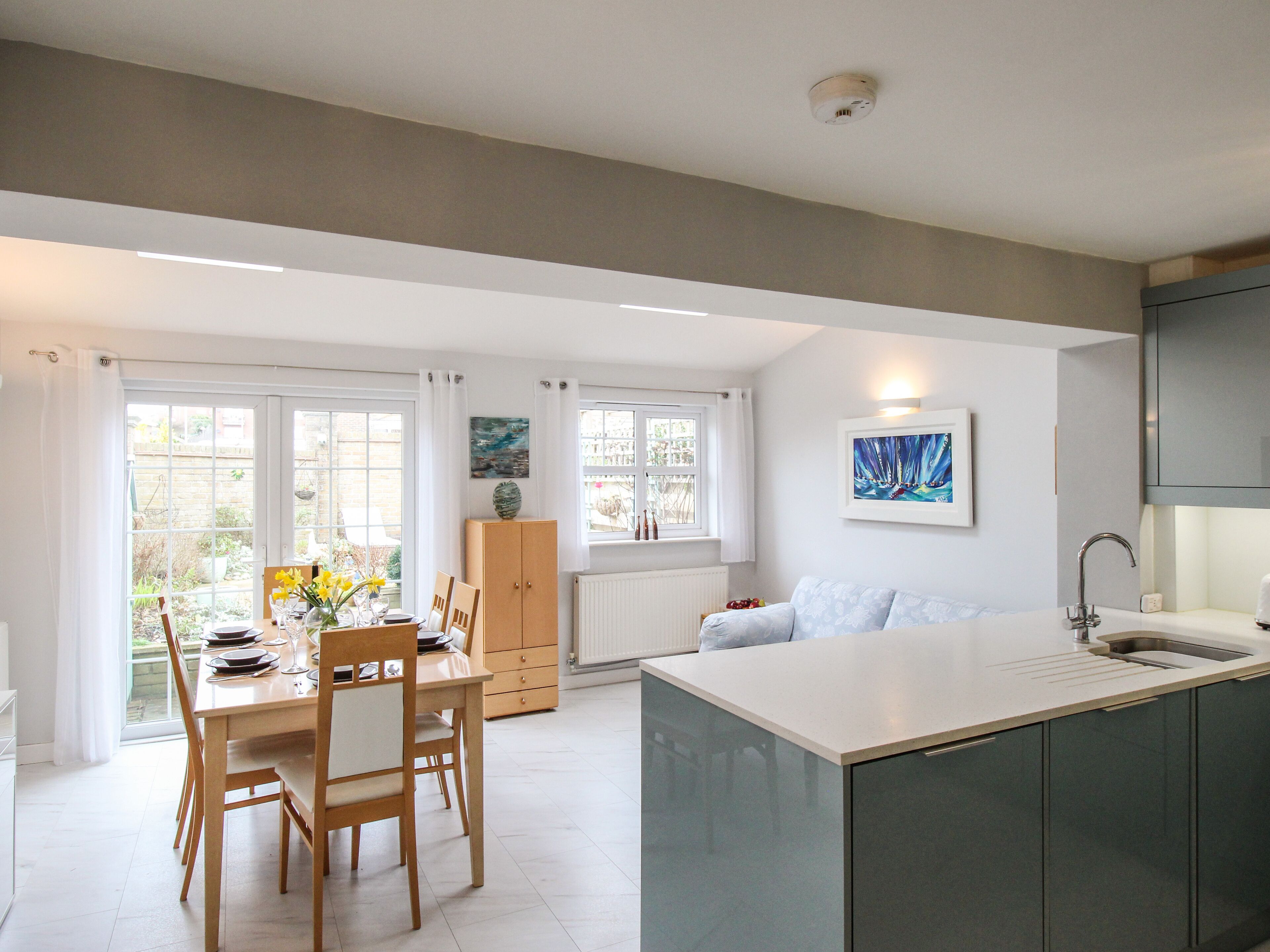 A modern kitchen with a central island, wooden dining table, and chairs. There is a window with curtains, a radiator, and a painting on the wall.