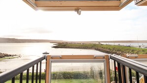 Une vue depuis un balcon avec une porte vitrée, donnant sur une étendue d’eau, une zone herbeuse et une côte lointaine.