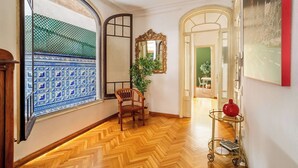 Un couloir doté d’un parquet en bois disposé en chevrons, d’une chaise en bois, d’un miroir et d’un vase rouge posé sur un chariot.