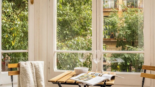 Une pièce avec une grande fenêtre, une table en bois sur laquelle repose un magazine, et deux chaises.