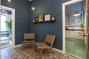 A room with two patterned chairs, a dark blue wall, a patterned floor, a kitchen with white cabinets, and a hanging light fixture. - The Collection - 4BD 3BTH with balcony in Eixample (Barcelona)