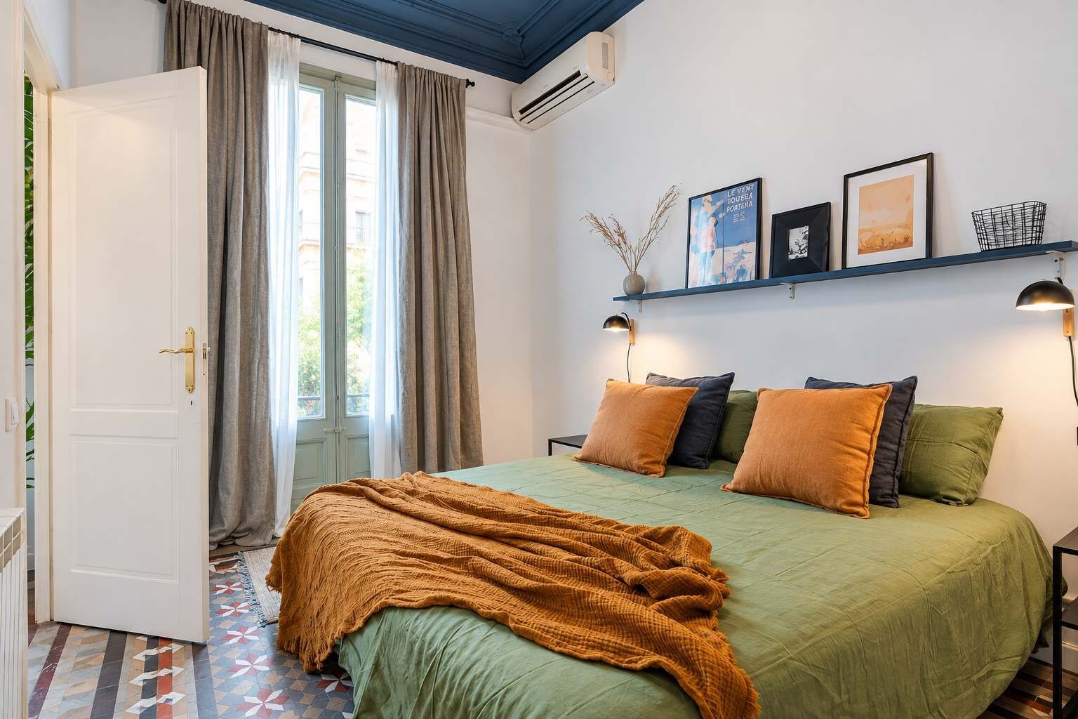 A bedroom with a bed, pillows, a blanket, a window with curtains, a wall-mounted shelf with pictures, and a door.