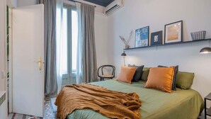 Ein Schlafzimmer mit Bett, einem Sessel, einem Regal mit Bildern und einem Fenster mit Vorhängen.
