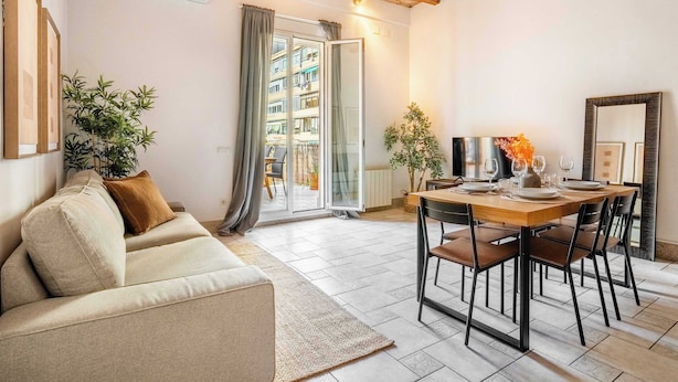 A modern living room with a sofa, dining table, and chairs. There is a large window with curtains, a plant, and a view of a building outside. - Historic apartment with private terrace in Example (Barcelona)