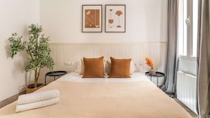 A hotel room with a large bed, two framed pictures on the wall, a potted plant, and a window with curtains. - Historic apartment with private terrace in Example (Barcelona)