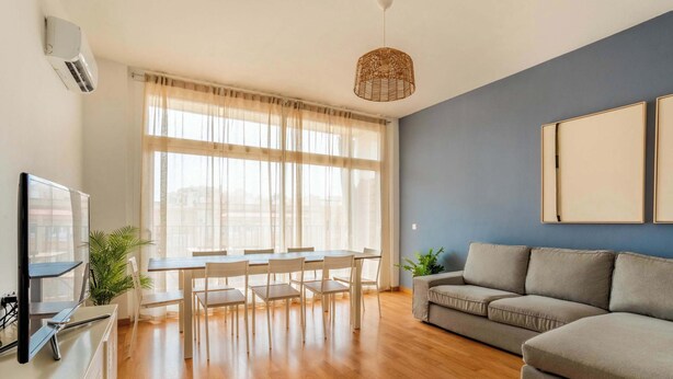 Un salon moderne avec un plancher en bois, un canapé gris, une table à manger avec des chaises, un téléviseur et une grande fenêtre ornée de rideaux légers.