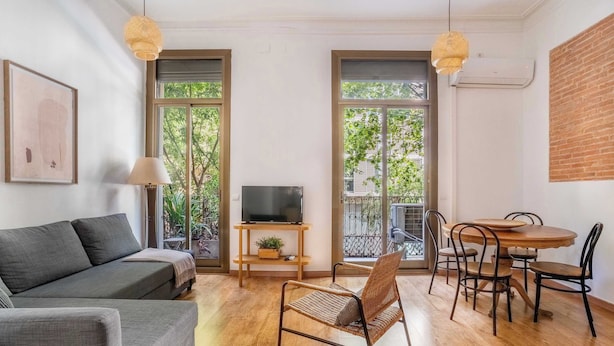 Ein modernes Wohnzimmer mit Sofa, Fernseher, Esstisch, Stühlen und großen Fenstern.