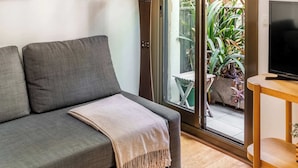 Ein modernes Wohnzimmer mit einem grauen Sofa, einer Lampe, einem Fernseher und einem großen Fenster mit Blick auf Grünflächen.