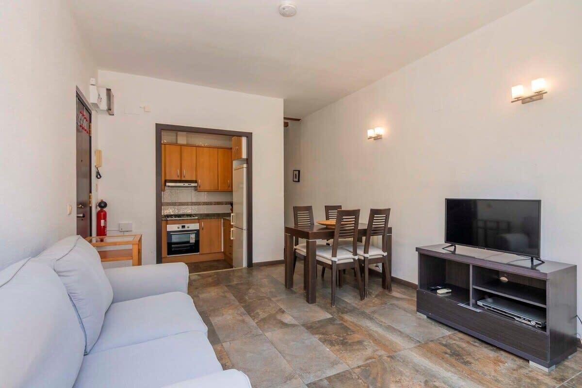 Una sala de estar con un sofá, una mesa de comedor con sillas, un televisor sobre un mueble bajo, y una cocina al fondo.