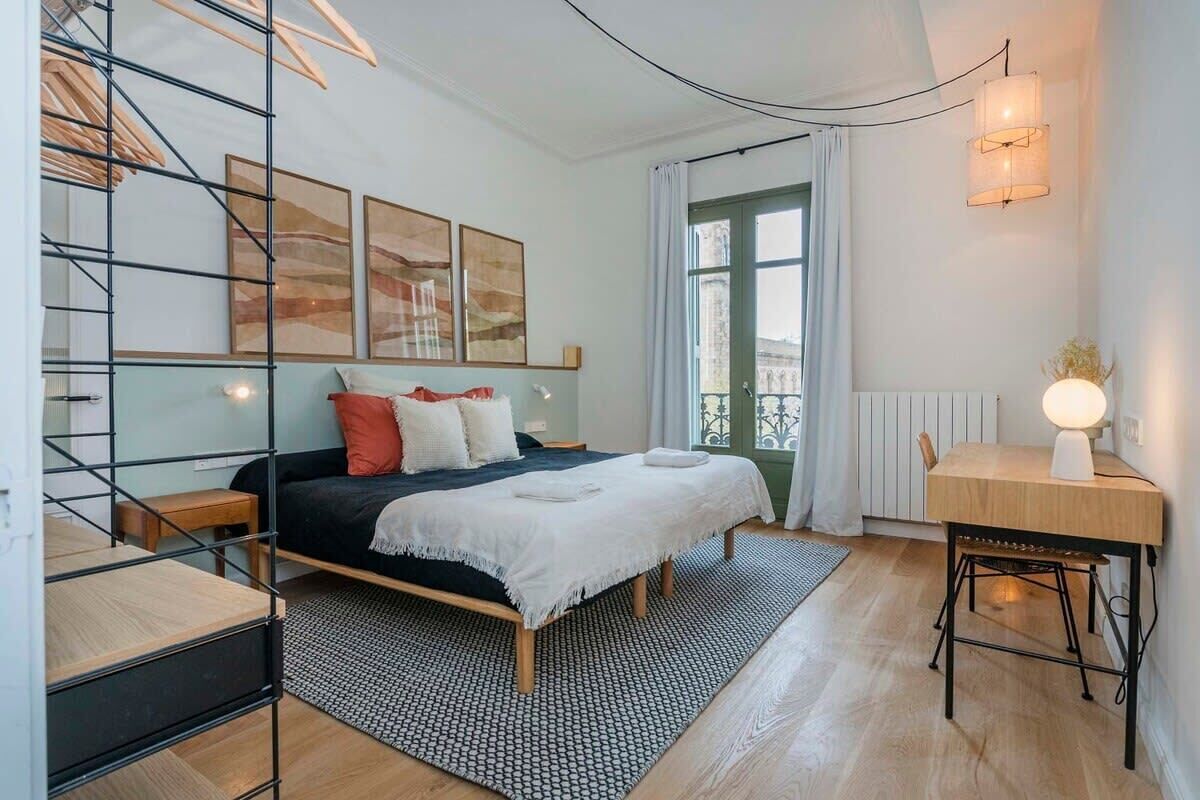 A bedroom with a bed, bedside tables, a desk, and a chair. There is a window with curtains, a radiator, and a wall-mounted lamp.