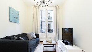 A modern living room with a black sofa, a coffee table, a flat-screen TV on a white stand, and a large window with curtains. - Centric 2bedroom in Las Ramblas (Barcelona)