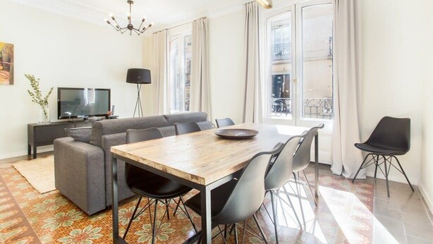A modern living room with a sofa, dining table, and chairs. There is a TV on a stand and a plant on the floor.