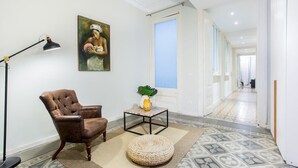 A room with a brown leather armchair, a small table with a plant, a woven ottoman, a painting on the wall, and a patterned floor.