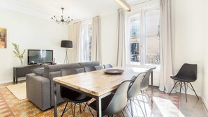 A modern living room with a sofa, a dining table, and chairs. There is a TV and a lamp.