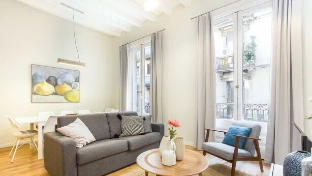 Un salon moderne avec un canapé gris, une table basse ronde et un fauteuil bleu. Un tableau orne le mur, et une fenêtre est agrémentée de rideaux.