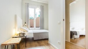 Une petite chambre bien éclairée, avec un lit, une table de chevet, une lampe et une vue sur un balcon.